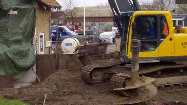En gravemaskine arbejder på en byggeplads, mens en lastbil og en betonblander er synlige i baggrunden.