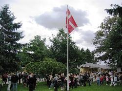 En stor gruppe mennesker samles under et dansk flag i en park med træer og bygninger i baggrunden.