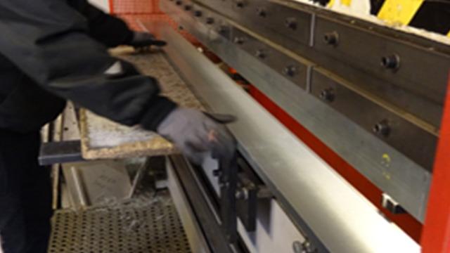 En person arbejder med en metalpresser, mens de justerer en metalplade med handsker på.