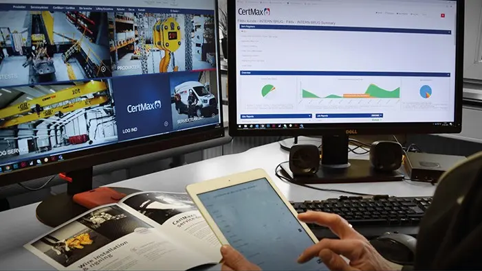 En person bruger en tablet, mens der er to computere med overvågningsbilleder og dataanalyse på skrivebordet.