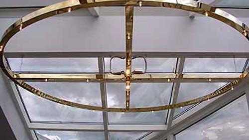 Moderne loftlampe i guld med en oval form, hængende under et glasloft med grå skyer i baggrunden.