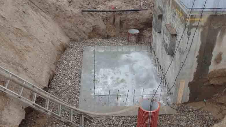 Byggeplads med en betonfundament, grus og armeringsjern i en udgravning ved en bygning.