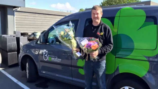En mand står foran en bil med blomster i hånden, smilende og glad.