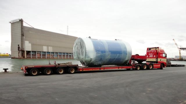 En stor lastbil transporterer en indpakket cylinder ved havnen, med en bygning i baggrunden.