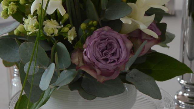 En smuk buket med lilla roser, hvide liljer og grønne blade i en hvid vase.
