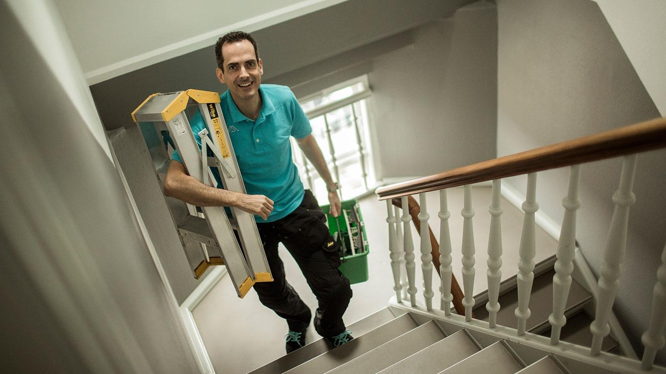 En håndværker bærer en stige og en kasse op ad trappen i et lyst hus.