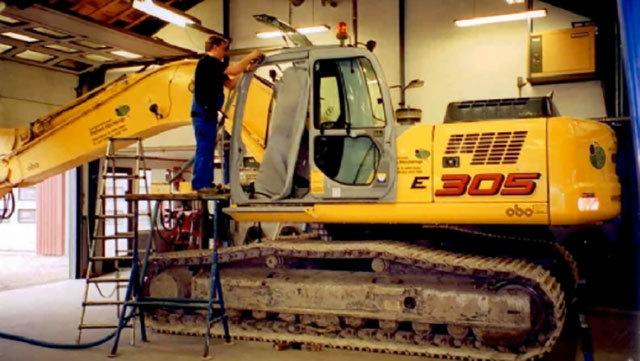 En mand arbejder på en gul gravemaskine E 305 i en værkstedshal med værktøj og stiger omkring.