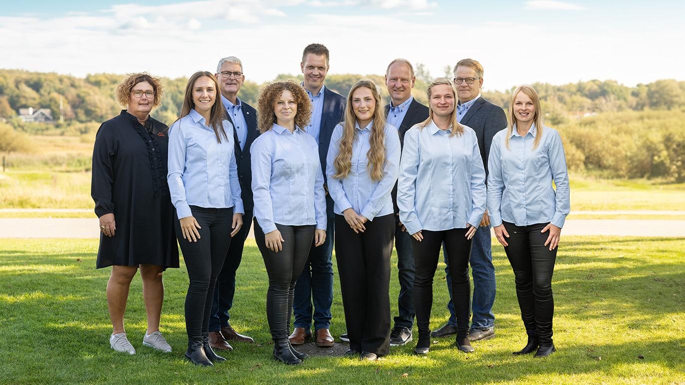 Gruppebillede af ti personer i skjorter, der står på en græsplæne med en naturskøn baggrund.