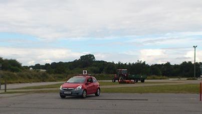 En rød bil kører på en parkeringsplads med en traktor i baggrunden under en skyet himmel.