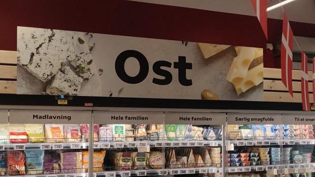 Ost afdeling i supermarked med forskellige oste og skilte, der viser ostetyper og madlavning.