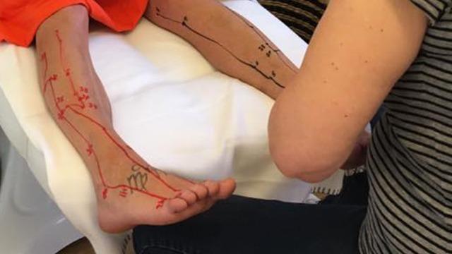 En person får lavet en tatovering med røde og sorte linjer på armene og fødderne, mens en anden person arbejder på designet.