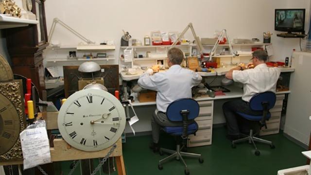 To personer arbejder på et urværksted med ure og værktøj på deres arbejdsborde.