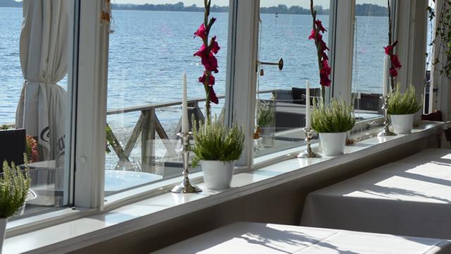 Udsigt fra en restaurant med vinduer mod vandet, dekoreret med blomster og grønne planter på bordene.