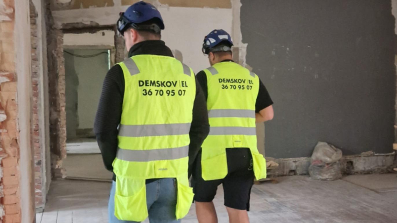 To personer i sikkerhedsveste går i et renoveret rum med synlige vægge og materialer.