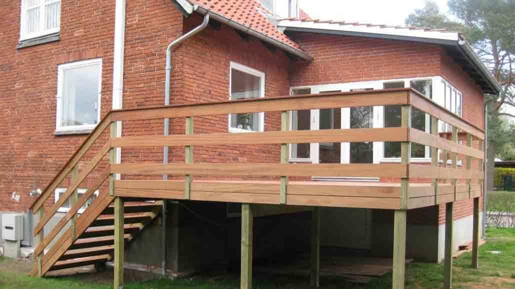 Hus med en træterrasse og trapper, beliggende i en have med græs og træer i baggrunden.