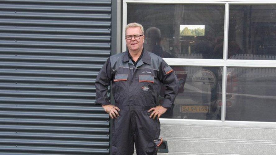 En mand i arbejdstøj står foran en garage med vinduer, iført briller og smilende.