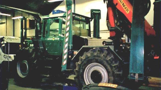 En grøn traktor med en kran står i en garage, omgivet af værktøj og dæk.