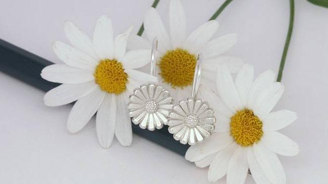 Sølvøreringe med blomsterdesign placeret ved siden af hvide margueritter.