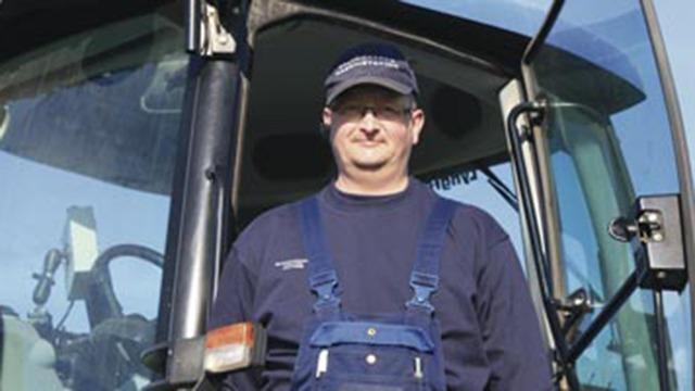 Landmand står i en traktor med blå overalls og kasket, klar til at arbejde på marken.