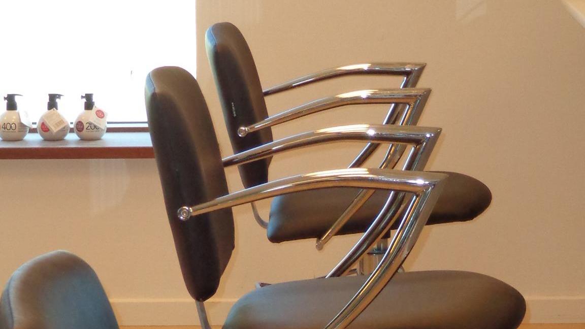 Fire frisørstole med metalrammer i en salon, med hårprodukter synlige i baggrunden.