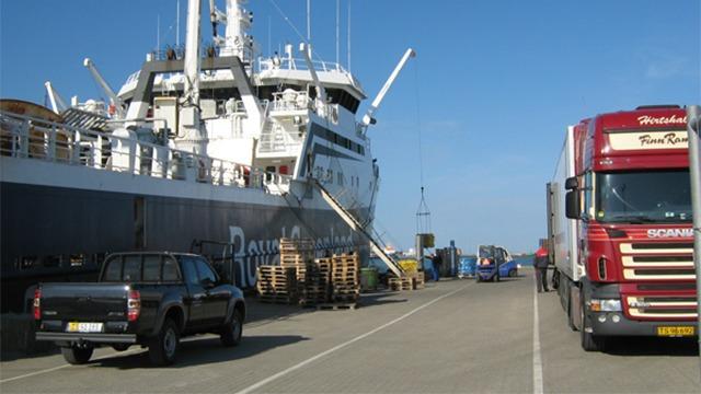 Lastbil og bil parkeret ved siden af et skib i havnen på en solrig dag.