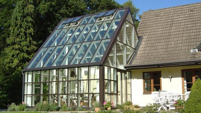 Et moderne glashus med skrå tag og store vinduer, omgivet af grønne træer og en solrig himmel.