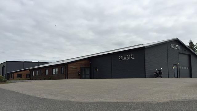 Moderne bygning med grå facade og trædetaljer, beliggende på en grusvej under en overskyet himmel.