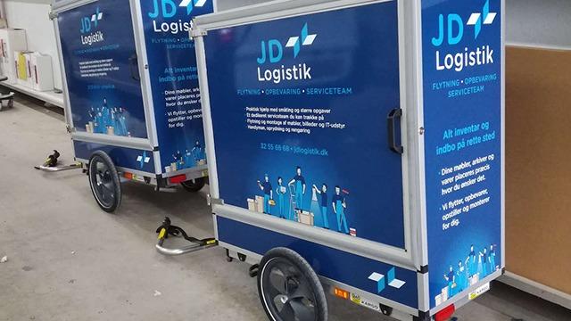 To vogne med JD Logistik logo og farver, der står i et lagerområde.