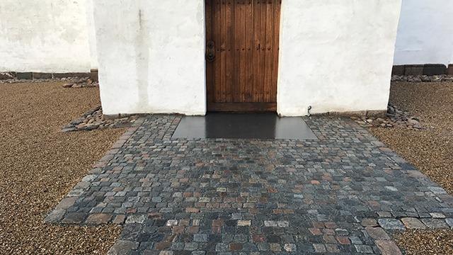 En trædør foran en belægning af brosten og grus, der fører ind i en hvid bygning.