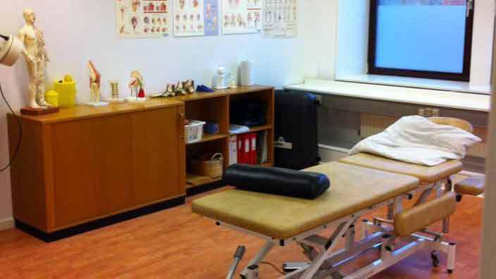 Behandlingsrum med en massagebriks, opbevaringsskabe og anatomiske plakater på væggen.