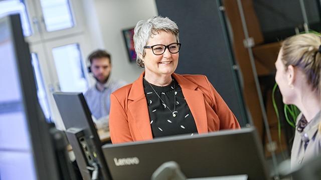 En smilende kvinde med gråt hår og briller sidder ved et skrivebord i et kontormiljø.