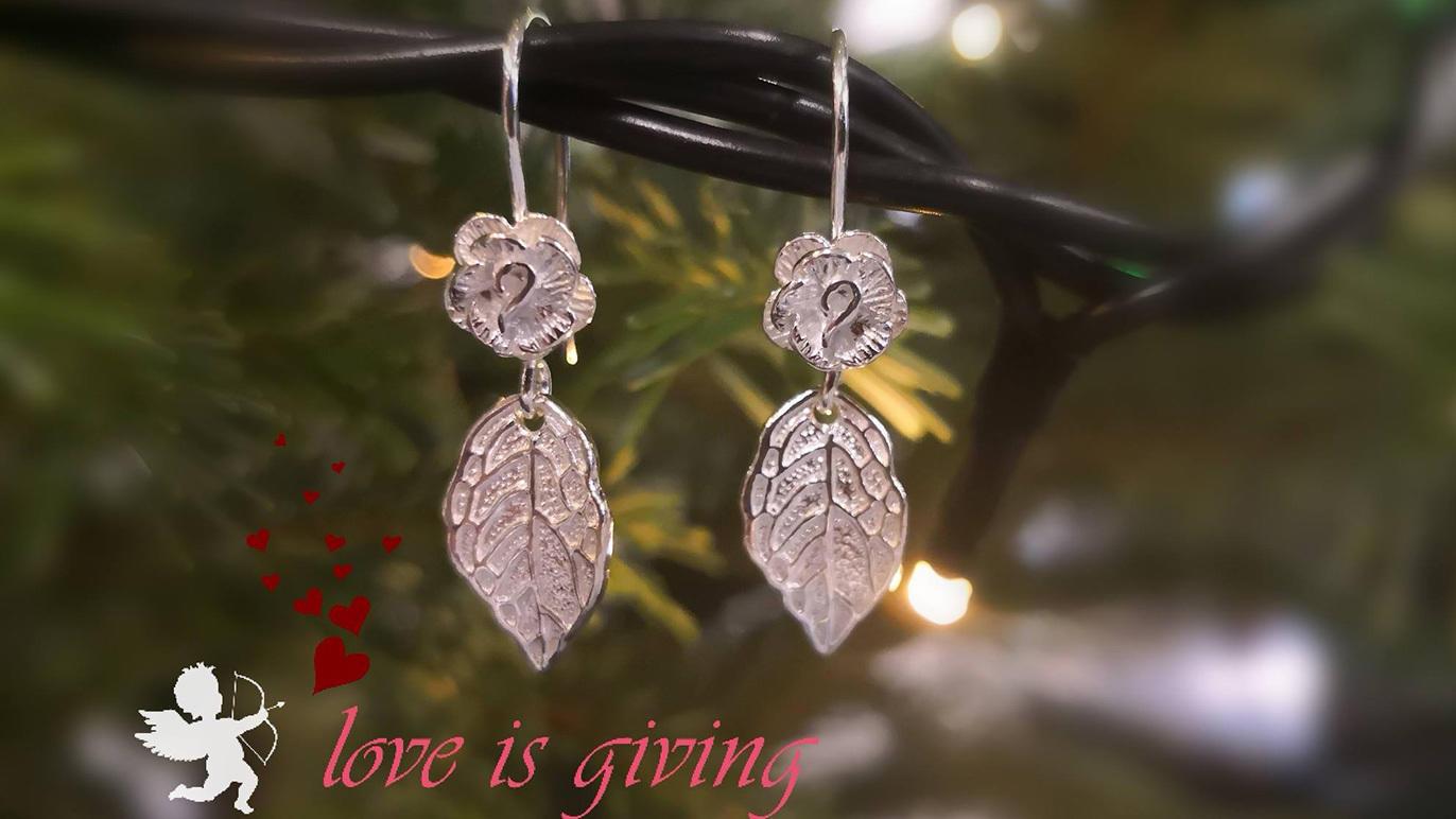 Sølvøreringe med bladdesign hængende på en gren, med teksten "love is giving" i bunden.