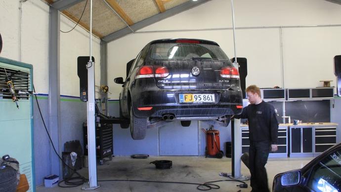 En mekaniker arbejder på en sort bil, der hænger oppe på en lift i et autoværksted.