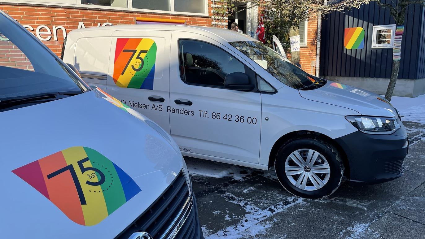 To biler med regnbuefarver og nummeret 75, parkeret foran en bygning med et logo.