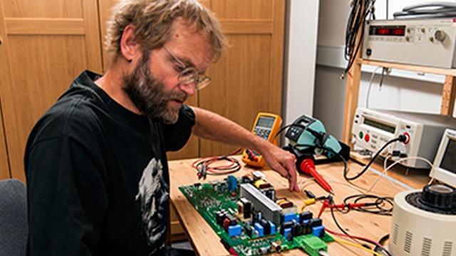 En mand arbejder med en kreds på et bord, omgivet af værktøj og måleinstrumenter i et laboratoriemiljø.