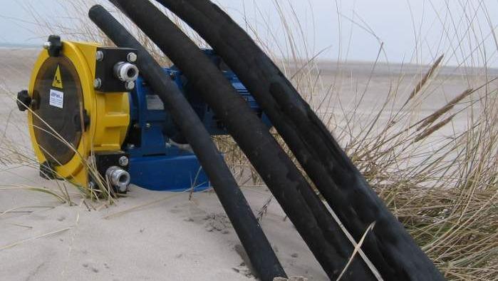 En blå og gul pumpe med sorte slanger ligger på sandet ved en strand med græs.