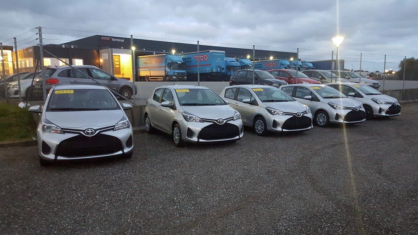 Flere sølvfarvede biler parkeret på en grusvej ved en bilforhandler under en overskyet himmel.