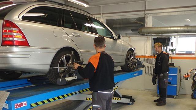 To biler bliver serviceret i et autoværksted, hvor mekanikere arbejder med hjuljustering.