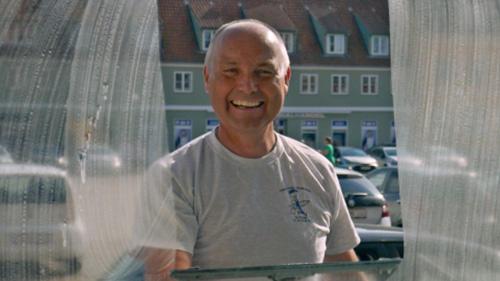 En glad mand står bag en plastikskærm med biler i baggrunden.