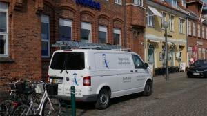 En hvid varevogn parkeret ved en bygning med mursten, cykler står parkeret i forgrunden.