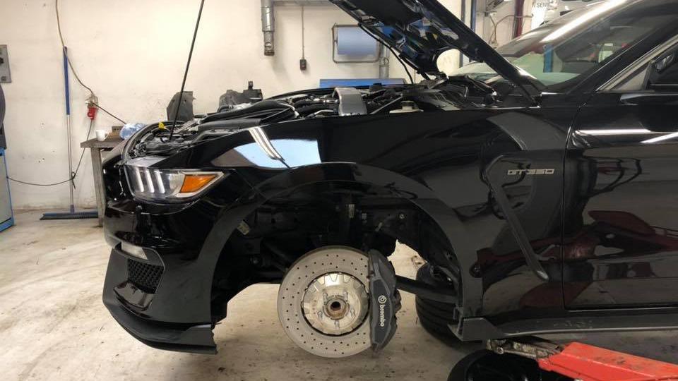Sort bil med åbent motorhjelm og afmonteret hjul i et værksted.