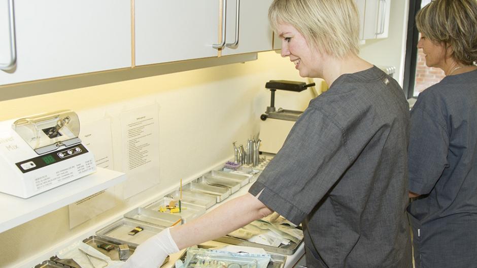 En kvinde i grå arbejdstøj organiserer medicinsk udstyr på en arbejdsplads i et sundhedsmiljø.