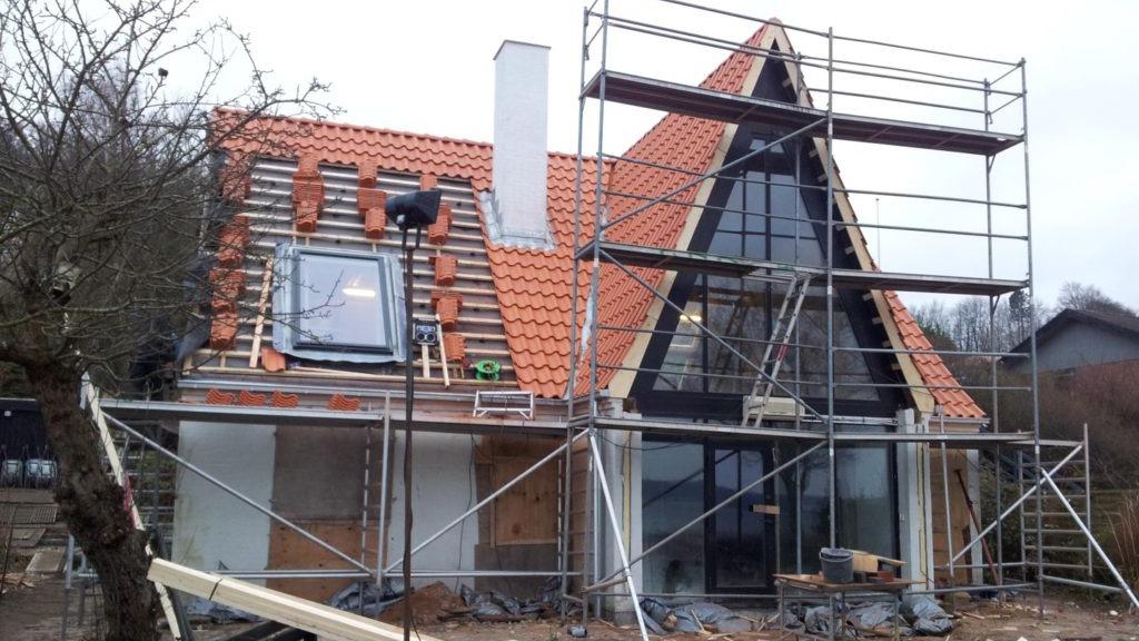 Hus under konstruktion med stillads, rødt tag og store vinduer i en landsby.