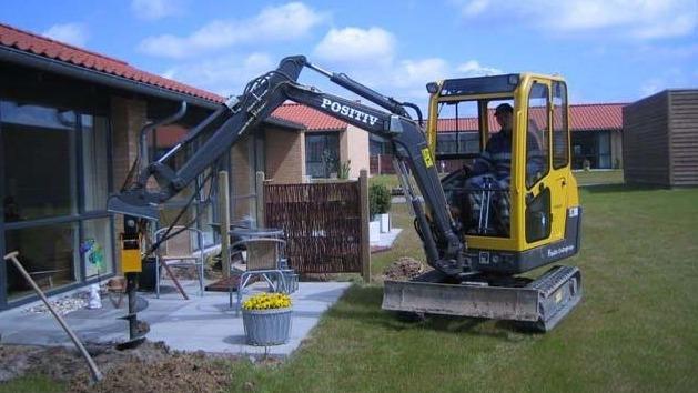 En gravemaskine arbejder på en græsplæne ved en bygning, med en person i førersædet og solskin i baggrunden.