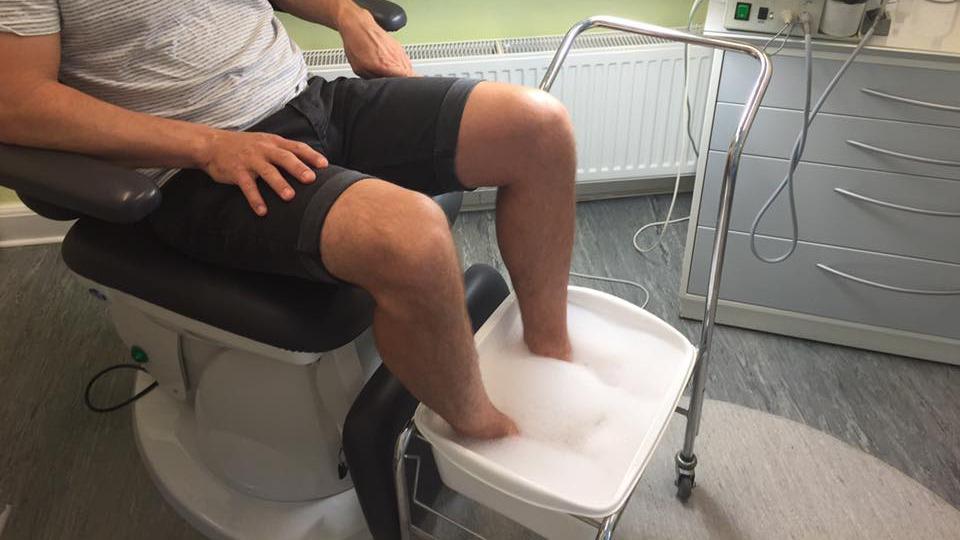 Mand sidder i en stol med fødderne i en skål med skum, mens han får behandling i en klinik.