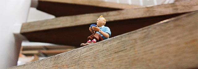En lille figur af Tintin sidder på en trætrappe med en baggrund af træ og indendørs miljø.