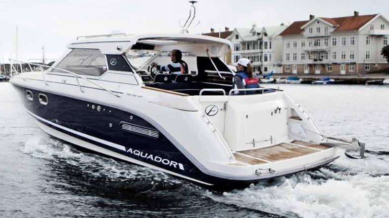 En motorbåd sejler gennem havnen med to personer ombord og bygninger i baggrunden.