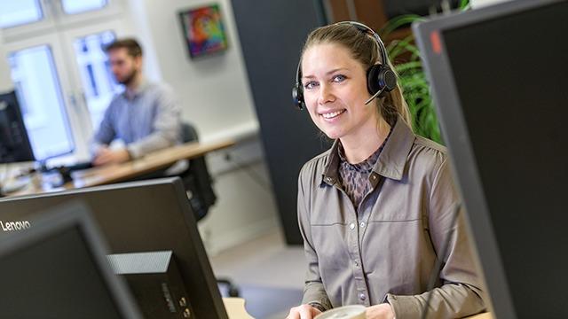 Kvindelig kundeservicemedarbejder med headset smiler ved skrivebordet i et kontormiljø.
