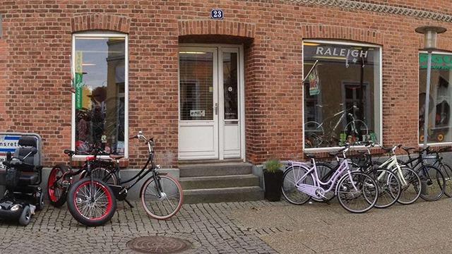 Cykelbutik med flere cykler parkeret foran en murstensbygning på en brostensbelagt gade.