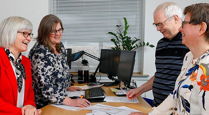 Fire personer står ved et skrivebord i et kontor, smilende og diskuterer dokumenter og computere.